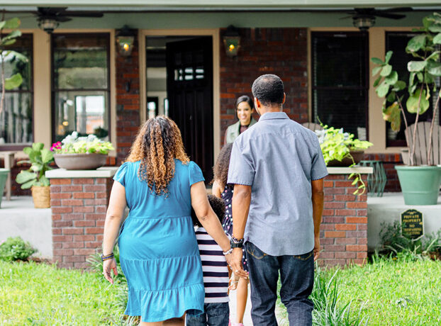 A couple walks hand in hand with their young child toward a smiling woman standing in front of a house with an open door